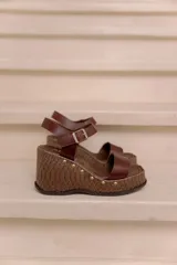 Sandalias de plataforma y taco alto confeccionadas en cuero vacuno marrón, con tira ancha en el empeine, tira ajustable con hebilla en el tobillo y detalles de tachas decorativas en la base.