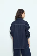 Sobrecamisa de jean color azul oscuro con estampado de rayas verticales blancas. Presenta un corte relajado, cuello clásico, cierre frontal con botones metálicos, mangas largas y dos bolsillos frontales de parche.