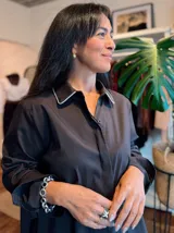 Camisa negra de corte holgado con cuello clásico decorado con una fina línea de strass brillante. Presenta cierre frontal con botones ocultos y mangas largas.