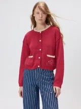 Cardigan de algodón grueso color rojo, con cuello redondo, cierre frontal de botones grandes y bolsillos cuadrados en el frente. Presenta detalles de pelo sintético en el cuello y los bolsillos, y logo bordado en uno de los bolsillos.