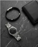 Pulsera doble de color negro, compuesta por una banda de cuerina trenzada y una placa metálica central con diseño de guarda griega grabada.