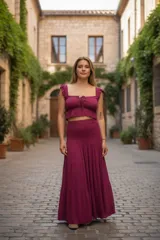 Conjunto de dos piezas en color uva, compuesto por un top corto y una pollera larga. El top cuenta con escote cuadrado, detalle de lazo frontal, breteles con volados y bajo elástico. La pollera es de largo maxi con diseño escalonado y cintura ancha elastizada tipo nido de abeja.