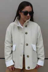 Campera de paño de lana en color beige jaspeado, con diseño oversize. Presenta detalles en pana color marrón chocolate en los puños y en las tapas de los bolsillos frontales. Cuenta con cierre frontal de botones grandes y cuello alto.