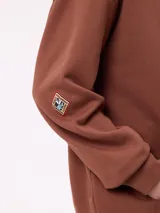 Canguro marrón con capucha y bolsillo frontal, con un diseño gráfico colorido en la espalda que representa una casa surrealista con el texto 'Supreal State of Mind'.