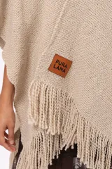 Ruana de tejido de punto en color arena, con diseño cruzado, escote en V y terminaciones con flecos en el ruedo. Incluye un parche de cuero con la marca Pura Lana en la parte frontal.