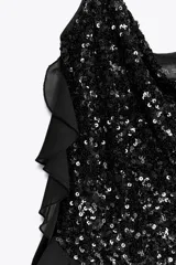 Vestido corto de escote fluido y tirantes finos, completamente cubierto de lentejuelas negras. Presenta detalles de volantes en tejido combinado que recorren el lateral y el frente de la prenda.