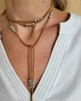 Collar estilo corbatín confeccionado en tiento de cuero color suela, con detalle de cuentas metálicas plateadas distribuidas a lo largo de las tiras.