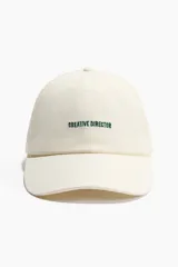 Gorra de sarga de algodón en color blanco, con diseño bordado frontal con la frase "Creative Director" en color verde y visera curvada.