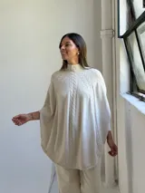 Poncho de tejido de punto color beige con cuello alto y diseño de trenzas verticales en el frente. Presenta mangas integradas con puños acanalados y un corte holgado con ruedo redondeado.
