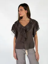 Blusa de color marrón oscuro, con escote en V y volados en cascada que rodean el cuello y descienden por el frente. Las mangas son cortas y amplias, con terminación en volados.