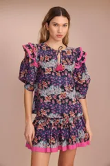 Blusa de algodón con estampado floral en tonos azul, rosa y naranja. Tiene cuello redondo con lazo y mangas cortas abullonadas con volados rosas.
