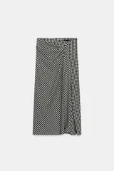 Falda midi de tiro alto con estampado de cuadros vichy en blanco y negro. Presenta un detalle de tejido drapeado en la parte delantera, bajo con abertura lateral y cierre lateral con cremallera oculta.