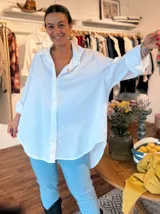 Camisa blanca de corte oversize, con cuello clásico, cierre frontal de botones ocultos y mangas largas con puños simples. Presenta un diseño de bajo redondeado con la parte trasera ligeramente más larga.