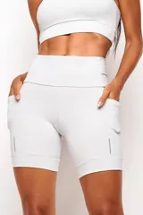Short biker deportivo de tiro alto, color blanco, con bolsillos laterales funcionales y detalles reflectivos en el ruedo. Confeccionado en tejido de alta performance con cintura anatómica y bolsillo interno 360°.