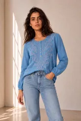 Blusa blanca de manga larga con detalles bordados en tono marrón en el pecho y las mangas, cuello redondo y cierre frontal con pequeños botones.