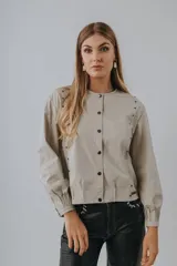 Campera bomber de cuero color beige, con cuello redondo, cierre frontal de botones a presión, mangas largas con puños fruncidos y detalles de tachas metálicas en los hombros y sisas.