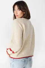 Sweater de punto color crudo con diseño de corazones rojos en las mangas, cuello redondo y terminaciones acanaladas en contraste rojo.