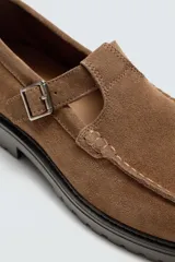 Zapato tipo Mary Jane de hombre, color marrón, fabricado en piel con acabado serraje (gamuza). Presenta cierre con hebilla lateral, detalle de bordón en el empeine y suela gruesa con volumen.