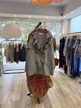 Gabardina de corte clásico en color verde militar, con solapas amplias y puños con detalles en estampado de cuadros escoceses en tonos azul, blanco y negro. Presenta cierre frontal cruzado con botones y bolsillos laterales.