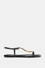 Sandalia plana de color negro con textura efecto piel de serpiente. Presenta una tira central con un aplique metálico dorado curvo y cierre de pulsera con hebilla ajustable en el tobillo.
