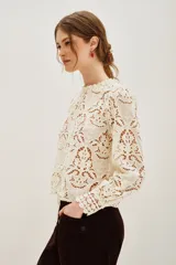 Camisa de encaje negro con cuello alto y mangas largas abullonadas. Presenta un diseño calado con motivos florales y cierre posterior.