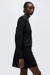 Vestido corto negro de sarga con escote redondo, hombreras finas y mangas largas con abertura en los puños. Presenta cintura entallada con pinzas, bolsillos ribeteados con solapa en el frente y cierre oculto en la parte trasera. Forrado.