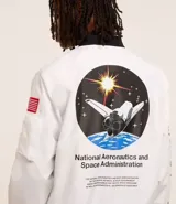 Campera tipo bomber blanca con detalles en negro, inspirada en uniformes espaciales de la NASA. Presenta parches con el logo de la NASA, la bandera de EE. UU. y texto en la manga que dice "Kennedy Space Center".