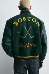 Cazadora estilo varsity con cuerpo de mezcla de lana en color verde oscuro y mangas de efecto piel a tono. Presenta cuello solapa, cierre frontal con botones a presión, bolsillos laterales de vivo y puños y bajo acanalados con rayas amarillas. Incluye bordados y parches decorativos en el frente con la inscripción Boston Archive.