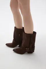 Bota corta de gamuza color chocolate, con punta fina y taco bajo. Presenta detalle de flecos en el lateral exterior y dos correas decorativas con hebillas metálicas en el tobillo.
