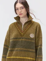 Pullover de punto con textura aterciopelada, diseño oversize, cuello polo con cierre frontal parcial y detalles de pespuntes en contraste. Presenta rayas horizontales en tonos tierra y un parche bordado con la letra M en el pecho.