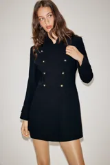 Vestido tipo blazer de corte estructurado, color negro, con cuello subido y cierre frontal cruzado con botones metálicos dorados. Presenta mangas largas con trabillas en los puños y bolsillos de vivo decorativos en el frente.