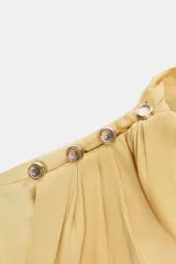 Blusa de manga larga color amarillo pálido, con cuello alto drapeado y detalle de botones dorados en el hombro izquierdo. Las mangas terminan en puños anchos.