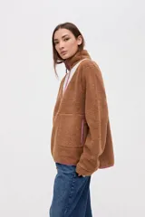 Buzo de sherpa color marrón con diseño de medio cierre frontal. Presenta un panel superior en forma de V en color beige y detalles de vivos en color fucsia en el cierre, cuello y bolsillos laterales. Cuenta con cuello alto y bolsillos frontales.