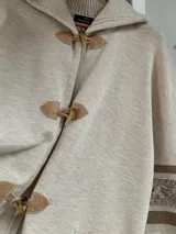 Saco tipo capa color beige, tejido en punto fino de lana con cierre de tancas de madera. Presenta cuello polo tejido y mangas anchas con detalles de trenzas y cintas tejidas.