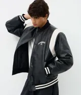 Campera bomber de poliuretano (PU) color negro con mangas en contraste de color blanco. Presenta cuello, puños y dobladillo elasticados con rayas blancas y negras, cierre frontal con botones a presión ocultos y bordado con la palabra 'Authentic 86' en el pecho.