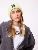 Gorro tipo beanie de punto acanalado en color lila, con parche rectangular negro en el frente que incluye la inscripción 'EAT THE WAVE'.