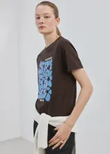 Camiseta marrón de algodón con cuello redondo y manga corta, con estampa frontal azul de flores y texto "Bloom More Worry Less".