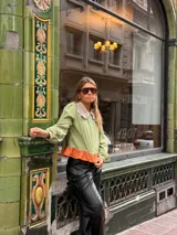 Camisa de manga larga en color verde oliva, con cuello babero en tono contrastante y detalle de volado en el ruedo en color naranja. Presenta cierre frontal con botones y mangas abullonadas con puños ajustados.