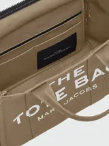 Cartera tipo tote bag de Marc Jacobs, confeccionada en lona color beige con el logo estampado en blanco al frente. Cuenta con doble asa superior, correa de hombro ajustable y extraíble, y cierre superior con cremallera.