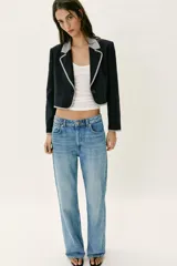 Blazer corta de color negro con cuello solapa, manga larga y hombreras. Presenta detalles combinados con estampado de cuadro vichy en el cuello, los bordes de la solapa, el ruedo y los puños. Cuenta con cierre frontal de un botón y falsos bolsillos delanteros de vivo.