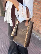 Bolso matero de cuero color chocolate con textura croco, asas cortas y detalle de costuras en los laterales. Incluye un dije con flecos en tonos beige y negro.