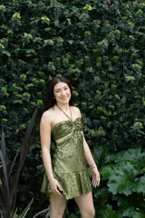 Vestido corto de lentejuelas color ciruela o morado oscuro, con escote corazón y detalle de argolla central que conecta con tirantes finos que atan al cuello. Presenta aberturas laterales (cut-outs) en la zona de la cintura y falda corta con vuelo.