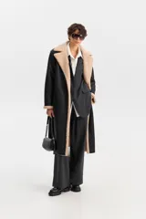 Abrigo largo de color negro con interior de corderito en contraste beige. Presenta un diseño de cierre cruzado con botones, solapas anchas y bolsillos laterales.