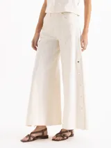 Pantalón de denim de algodón pima color blanco off-white, de tiro alto y silueta amplia de corte ancho. Presenta costuras verticales visibles y broches metálicos decorativos y funcionales en los laterales.