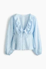 Blusa celeste con estampado de lunares blancos, escote en V pronunciado con volados, mangas largas abullonadas con puños elásticos y lazo ajustable en el frente.