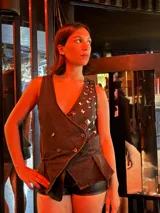 Chaleco sastrero de denim negro con escote en V profundo, cierre cruzado con doble abotonadura, tachas metálicas aplicadas en el frente y ruedo irregular con volados.