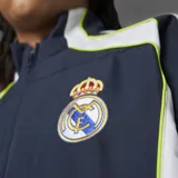 Campera deportiva Adidas del Real Madrid, color azul marino con detalles en blanco y amarillo. Presenta cierre frontal, cuello alto, bolsillos laterales, puños elásticos, dobladillo ajustable y escudo del club bordado en el pecho.