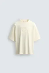 Camiseta de manga corta y cuello redondo, color crema o blanco roto, con un parche texturizado en el pecho que simula barras verticales.