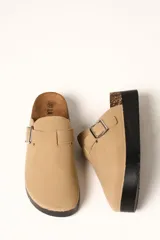 Zueco de plataforma color beige, con capellada de gamuza sintética, detalle de hebilla metálica lateral y suela de goma negra con detalle de corcho en el talón.
