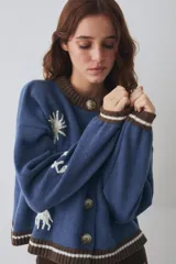 Cardigan de lana color crema con cuello redondo, cierre frontal de botones y mangas largas con puños ajustados. Presenta bordados decorativos en el frente que incluyen un sol, olas y estrellas, con detalles en azul en el cuello y el ruedo.